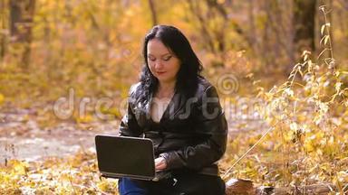 一个年轻的黑发女人在阳光明媚的秋日坐在公园里，手提电脑远程工作